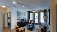 This single-floor residence offers a thoughtfully designed living area that combines contemporary style with functional comfort. The main living space is anchored by a striking large-scale abstract artwork in muted blue and gray tones, mounted above a sleek, dark credenza that serves as a media console and display surface. Track lighting runs along the ceiling, providing adjustable, directional illumination that complements the ceiling fan with an integrated light fixture positioned at the room's center.

The space benefits from generous natural light, courtesy of tall floor-to-ceiling windows and glass doors dressed with dark curtain panels and horizontal blinds. These openings offer views of the surrounding exterior and lend an open, airy quality to the interior. Light-toned walls and light hardwood-style flooring enhance the brightness throughout.

The seating arrangement features a neutral sofa, cognac leather accent chairs with a modern frame, and a dark rectangular coffee table, all anchored by a patterned area rug. The overall ambiance is modern and sophisticated, with a carefully curated mix of textures and tones that create an inviting yet polished atmosphere.