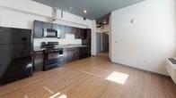 This single-floor unit offers a well-organized layout that balances functionality and comfort. The kitchen is a standout feature, equipped with rich dark espresso cabinetry, black appliances including a refrigerator, electric range, built-in microwave, and dishwasher, along with a stainless steel sink and ample counter space. Exposed ductwork along the ceiling adds an industrial character to the space. Warm wood-tone laminate flooring runs throughout the unit, creating visual continuity between rooms. The open layout connects the kitchen to the living area, offering a flexible and spacious feel. The bedroom receives generous natural light through multiple windows, including a higher transom-style window, and is equipped with a wall-mounted PTAC heating and cooling unit for individual climate control. The bathroom is clean and well-appointed, featuring a full soaking tub with a tiled surround, built-in shelving for storage, and matching wood-tone flooring. The overall aesthetic is modern and minimal, with neutral white walls contrasting against the dark cabinetry and warm flooring, creating a cohesive and inviting environment throughout.