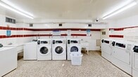 Located at 120 Shelborne Ave in Toronto, this shared laundry facility is a well-equipped, single-floor space designed to serve building residents efficiently. The room is generously sized, providing ample floor space for comfortable movement between machines and laundry tasks.

The interior features white ceramic wall tiles accented with a bold red horizontal stripe running along the perimeter, giving the room a clean and distinct visual character. The terrazzo flooring throughout is durable and easy to maintain, well-suited to the demands of a high-use communal facility.

The room is outfitted with multiple front-loading washing machines and a separate row of top-loading washers, alongside a full bank of dryers — all commercial-grade white units equipped with digital controls. A utility sink is available for hand-washing or pre-treating garments, and a dedicated folding table with seating provides a practical workspace for residents.

Bright fluorescent ceiling fixtures ensure the entire space is well-lit, while informational signage and a bulletin board keep residents informed of laundry guidelines and building notices. A security camera is also present, contributing to the safety of the space. The room includes a utility door and is thoughtfully laid out to maximize usability and flow throughout the facility.