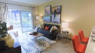 This single-floor space is designed with comfort and contemporary style in mind. The living area serves as the heart of the home, showcasing warm yellow-toned walls that create an inviting and cheerful ambiance. Light hardwood flooring runs throughout, complementing the overall modern aesthetic.

The living room is thoughtfully arranged around a charcoal gray sofa accented with decorative pillows in neutral and warm tones. A patterned area rug anchors the seating area, while a wooden coffee table and sleek metallic nesting side tables add functional elegance. Two table lamps provide warm ambient lighting, enhancing the cozy atmosphere.

A gallery wall of bold, colorful artwork featuring urban and lifestyle themes adds a dynamic focal point to the space. Sliding glass doors dressed with patterned curtains flood the room with natural light and open directly to an outdoor patio area, offering a seamless connection between indoor and outdoor living. The view through the glass doors reveals lush greenery, adding a touch of nature to the interior.

Overall, the space strikes a balance between modern design and relaxed livability, making it ideal for both everyday living and entertaining.
