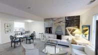 This two-floor property offers a thoughtfully arranged interior centered around an open-plan living and dining area. The living space is anchored by a generously sized white sofa ensemble, complemented by a glass-top coffee table and light-toned carpeting that extends throughout the room, creating a cohesive, understated aesthetic. A striking feature of the space is the large-scale, abstract artwork displayed prominently on the main wall, introducing dynamic color and artistic character to an otherwise neutral palette. The wall treatment includes what appears to be a fireplace surround integrated into the artwork display, adding an architectural focal point to the room. Adjacent to the living area, a dining space is furnished with black metal chairs around a glass-topped table, maintaining the space's clean, contemporary feel. The kitchen is partially visible beyond the dining area, suggesting an open and connected layout. White walls and a textured ceiling help to maximize the sense of light and openness throughout. The overall ambiance is modern and gallery-like, with the bold artwork serving as the defining design element of the main living level.