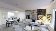This two-floor property offers a thoughtfully arranged interior centered around an open-plan living and dining area. The living space is anchored by a generously sized white sofa ensemble, complemented by a glass-top coffee table and light-toned carpeting that extends throughout the room, creating a cohesive, understated aesthetic. A striking feature of the space is the large-scale, abstract artwork displayed prominently on the main wall, introducing dynamic color and artistic character to an otherwise neutral palette. The wall treatment includes what appears to be a fireplace surround integrated into the artwork display, adding an architectural focal point to the room. Adjacent to the living area, a dining space is furnished with black metal chairs around a glass-topped table, maintaining the space's clean, contemporary feel. The kitchen is partially visible beyond the dining area, suggesting an open and connected layout. White walls and a textured ceiling help to maximize the sense of light and openness throughout. The overall ambiance is modern and gallery-like, with the bold artwork serving as the defining design element of the main living level.