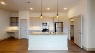 This single-floor residence showcases a clean, contemporary design with a cohesive neutral palette throughout. The open-concept kitchen serves as the centerpiece of the living area, anchored by a large white island with a granite countertop and integrated sink. Two geometric gold pendant lights hang above the island, adding a decorative accent against the recessed ceiling lighting. The kitchen is equipped with stainless steel appliances, including a refrigerator, range, and built-in microwave, all set against soft gray shaker-style cabinetry and a white subway tile backsplash. Wood-look luxury vinyl flooring runs continuously through the main living areas, lending warmth and visual continuity.

The entry foyer is clean and uncluttered, with white walls and recessed lighting that establish a bright, welcoming introduction to the space. Additional rooms feature the same flooring and crisp white walls with panel-style doors. The bathroom is finished with marble-look tile flooring and wall surround, a granite-top vanity with gray cabinetry, and gold-tone fixtures. A walk-in closet with built-in shelving and multiple hanging rods provides generous, well-organized storage. Overall, the space presents a fresh, move-in-ready aesthetic with consistent, quality finishes throughout.