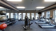 This fitness center is thoughtfully designed to provide a comprehensive workout environment for users of all fitness levels. The space is anchored by a generous selection of professional-grade equipment, including treadmills, an elliptical, cable machines, and a full dumbbell rack, offering a well-rounded range of strength and cardio training options. Large windows line the perimeter of the room, flooding the space with natural light while framing pleasant outdoor views of the surrounding greenery, creating an energizing and open atmosphere. Floor-to-ceiling mirrors along one wall enhance the sense of depth and allow users to monitor their form during workouts. The ceiling features recessed panel lighting that supplements natural light, ensuring consistent, well-lit conditions at all hours. Crown molding details along the upper walls add a refined architectural touch that elevates the overall aesthetic of the room. Neutral-toned walls are complemented by durable, textured carpet flooring in a herringbone-style pattern, offering both practicality and visual interest. A wall-mounted television is conveniently positioned for viewing during workouts. The layout is spacious and thoughtfully organized, with clear zones for cardio and strength training, making the space both functional and comfortable to navigate.