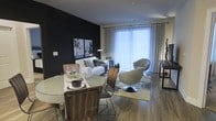 This single-floor residence showcases a striking contemporary design that makes a lasting impression. The open-concept living and dining area is anchored by a bold black accent wall, which serves as a dramatic backdrop for curated wall art and a clean-lined white sofa. The space is flooded with natural light streaming through expansive floor-to-ceiling windows dressed in sheer curtains, creating an airy, luminous ambiance that complements the rich hardwood flooring throughout.

The dining area features a round table paired with wooden chairs, offering an intimate yet functional setting for gatherings. The living space flows naturally from the dining zone, highlighted by a statement swivel lounge chair and a soft area rug that grounds the seating arrangement. The contrast between the dark accent wall and the bright, light-filled windows creates a dynamic visual balance that defines the overall character of the space.

The thoughtful layout maximizes the sense of openness while maintaining distinct functional zones. Modern pendant lighting and carefully selected décor add layers of sophistication, making this a well-appointed space that is both visually compelling and highly livable.