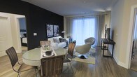 This single-floor residence showcases a striking contemporary design that makes a lasting impression. The open-concept living and dining area is anchored by a bold black accent wall, which serves as a dramatic backdrop for curated wall art and a clean-lined white sofa. The space is flooded with natural light streaming through expansive floor-to-ceiling windows dressed in sheer curtains, creating an airy, luminous ambiance that complements the rich hardwood flooring throughout.

The dining area features a round table paired with wooden chairs, offering an intimate yet functional setting for gatherings. The living space flows naturally from the dining zone, highlighted by a statement swivel lounge chair and a soft area rug that grounds the seating arrangement. The contrast between the dark accent wall and the bright, light-filled windows creates a dynamic visual balance that defines the overall character of the space.

The thoughtful layout maximizes the sense of openness while maintaining distinct functional zones. Modern pendant lighting and carefully selected décor add layers of sophistication, making this a well-appointed space that is both visually compelling and highly livable.