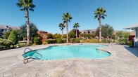 The Grand Palms pool facility is a well-appointed outdoor amenity space designed to evoke a resort-like atmosphere. At its centerpiece sits a generously sized freeform swimming pool with clear, blue-tinted water, framed by a broad stamped concrete deck ideal for lounging. Towering palm trees and mature landscaping surround the perimeter, providing both shade and a tropical aesthetic beneath open blue skies.

Adjacent to the pool, a substantial covered pavilion serves as the hub of the outdoor gathering area. The structure features a standing-seam metal roof supported by distinctive stacked-stone columns, lending the space a polished, upscale character. The pavilion floor is finished in large-format slate-style tile, continuing the refined aesthetic throughout. Comfortable lounge chairs are positioned under the covered area, offering shaded views of the pool. A built-in grill or outdoor kitchen station is also present, making the space well-suited for social gatherings.

The facility entrance is marked by arched architectural details, large picture windows, and stone accent columns that echo the pavilion's design language. The cohesive use of natural stone, neutral siding, and decorative tile throughout creates a unified, inviting ambiance. Brick residential buildings are visible in the background, providing context for the communal nature of this outdoor space.