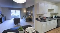 This single-floor residence offers a thoughtfully designed open-concept layout that seamlessly connects the kitchen, dining, and living areas into one cohesive living space. The kitchen features crisp white cabinetry with raised-panel doors, dark granite-style countertops, and ample storage, creating a clean and functional workspace. It flows naturally into the dining area, where a dark-toned table set with decorative place settings adds an elegant touch, complemented by a contemporary overhead light fixture.

The living room anchors the space with a soft, neutral color palette, featuring a charcoal accent wall that adds depth and contrast against the lighter surroundings. Plush carpeting runs throughout the living area, contributing to a warm and comfortable ambiance. A large sliding glass door or wide window at the far end of the living room floods the space with natural light and offers views of lush greenery outside. A sofa with coordinating accent pillows and a floor lamp complete the cozy yet refined atmosphere. A hallway visible from the living area suggests additional private spaces beyond, making this a well-rounded and functional residence.