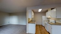 This single-floor space offers a practical and functional layout centered around an open-concept design that connects the kitchen and living area seamlessly. The kitchen is well-equipped with white cabinetry providing ample storage, a stainless steel sink set into a countertop peninsula, a white refrigerator, and a white electric range with overhead microwave. A skylight-style ceiling fixture brightens the kitchen, enhancing its clean, utilitarian aesthetic. The hardwood flooring in the kitchen transitions into plush carpeting in the adjacent living area, creating a clear distinction between the two zones while maintaining an open, airy feel. The living area is spacious and features neutral-toned walls and a textured ceiling, lending the space a calm and understated character. The open passthrough between the kitchen peninsula and the living room encourages connectivity and ease of movement throughout the space. Overall, the layout is efficiently designed to maximize usability, making it well-suited for everyday living. The neutral color palette throughout — from the walls to the cabinetry — provides a versatile backdrop that can easily be personalized to suit a variety of interior styles.
