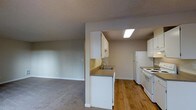 This single-floor space offers a practical and functional layout centered around an open-concept design that connects the kitchen and living area seamlessly. The kitchen is well-equipped with white cabinetry providing ample storage, a stainless steel sink set into a countertop peninsula, a white refrigerator, and a white electric range with overhead microwave. A skylight-style ceiling fixture brightens the kitchen, enhancing its clean, utilitarian aesthetic. The hardwood flooring in the kitchen transitions into plush carpeting in the adjacent living area, creating a clear distinction between the two zones while maintaining an open, airy feel. The living area is spacious and features neutral-toned walls and a textured ceiling, lending the space a calm and understated character. The open passthrough between the kitchen peninsula and the living room encourages connectivity and ease of movement throughout the space. Overall, the layout is efficiently designed to maximize usability, making it well-suited for everyday living. The neutral color palette throughout — from the walls to the cabinetry — provides a versatile backdrop that can easily be personalized to suit a variety of interior styles.
