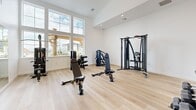 Located in Herriman, Utah, this dedicated fitness facility offers a comprehensive workout environment across two exercise rooms connected by a hallway. The space is thoughtfully designed with a clean, modern aesthetic, featuring crisp white walls, light hardwood flooring throughout, and recessed lighting that creates a bright and energizing atmosphere.

The first exercise room is generously sized and bathed in natural light from large windows that frame views of the surrounding Herriman neighborhood. This room is equipped with a full cable machine, adjustable weight benches, a dumbbell rack, and a variety of strength training machines, making it ideal for resistance and functional training. Soaring ceilings in this area amplify the sense of openness.

The second exercise room is dedicated to cardiovascular training and features a selection of cardio machines including treadmills, ellipticals, a recumbent bike, and a rowing machine. A row of wide windows lines the wall, filling the room with natural light. Full-length wall mirrors in the strength area enhance spatial depth and support form monitoring during workouts.

Together, the two rooms provide a well-rounded fitness experience suitable for a wide range of training styles, all within a clean, modern, and well-lit facility.