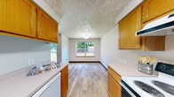 Located on Hidden Creek Loop NE in Keizer, Oregon, this single-floor home offers four bedrooms and one bathroom within a compact, efficiently laid-out floor plan. The galley-style kitchen is well-appointed with warm honey-toned oak cabinetry lining both walls, white appliances including an electric range and dishwasher, and ample counter space. A window at the end of the kitchen and dining area frames a view of the lush greenery outside, filling the space with natural light. The wood-look vinyl plank flooring runs throughout the kitchen and common areas, providing a clean and cohesive look, while a distinctive textured ceiling adds character to the living spaces. The full bathroom features the same wood-look flooring, an oak vanity with a white countertop, and a shower-tub combination. The four bedrooms are carpeted in a neutral tone and are naturally lit by large windows that overlook the surrounding greenery and neighborhood. The spacious primary bedroom offers generous room for furnishings. A hallway connects the bedrooms, creating a functional flow throughout the home. Warm wood baseboards and trim details unify the interior, giving the space a consistent and welcoming aesthetic.