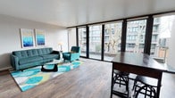 This contemporary multi-floor space is designed with an emphasis on openness, natural light, and modern living. The main living area makes an immediate impression with its expansive floor-to-ceiling windows and sliding glass doors that frame views of the surrounding urban landscape, including a mature tree and neighboring architecture. These large openings flood the interior with natural light while providing seamless access to a private balcony, blurring the line between indoor and outdoor living.

The living space features warm wood-tone flooring that runs throughout, complementing the clean white ceilings and dark window framing for a balanced, contemporary aesthetic. A bar-height counter with seating anchors one side of the room, creating a natural division between the living and dining or kitchen areas without closing off the open-concept layout.

The overall ambiance is light, airy, and modern, with a neutral palette punctuated by thoughtful design details. With two floors of living space, the property offers both social and private zones, making it well-suited for a variety of uses. The combination of generous glazing, quality finishes, and urban views creates a space that feels connected to its city surroundings while maintaining a comfortable and refined interior atmosphere.