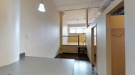 This single-floor space presents a distinctive industrial character, defined by its exposed ceiling infrastructure, including visible pipes and a track lighting system that runs across the ceiling. The open layout creates a sense of spaciousness, complemented by elevated ceilings that enhance the airy atmosphere throughout.

A striking wrought-iron spiral staircase serves as a notable architectural feature, positioned near a set of metal railings that define an upper landing area. This design element adds both visual interest and a sense of vertical dimension to the space. Natural light enters through a window on the far wall, brightening the interior and contributing to an open, welcoming feel.

The flooring throughout is a dark-toned material that contrasts with the light-colored walls, creating a balanced visual aesthetic. A pendant light fixture with a classic bell-shaped shade hangs prominently in the foreground, adding a touch of character. Additional doorways visible along one wall suggest further utility or storage areas. Overall, the space carries a raw, loft-like aesthetic suited to a variety of functional uses.