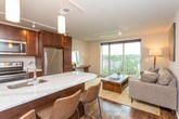 This well-appointed single-floor unit offers a thoughtfully designed layout that balances comfort and functionality. The living area serves as the heart of the space, featuring warm hardwood-style flooring, track lighting, and a large sliding glass door that floods the room with natural light and opens to a private balcony overlooking city views. A colorful canvas print adds a vibrant touch to the neutral-toned walls.

The galley-style kitchen is equipped with rich dark cabinetry, light stone countertops, stainless steel appliances including a refrigerator, electric range, built-in microwave, and dishwasher. Pendant lighting and track fixtures illuminate the workspace efficiently.

Both bedrooms feature plush carpeting, track lighting, and neutral walls that create a calm, restful atmosphere. Each room is complemented by warm wood-toned furniture and coordinated decor, lending a cohesive, mid-century modern aesthetic throughout.

The bathroom is finished with wood-look tile flooring, a dark vanity with a white countertop, a large mirror, and a sliding glass shower enclosure. Completing the unit is a dedicated laundry area with a stacked washer and dryer, offering the convenience of in-unit laundry alongside additional shelving for storage.