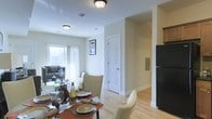 This two-story residence offers a thoughtfully designed layout that blends comfort and functionality across both levels. The main floor showcases an open-concept living and dining area bathed in natural light from large windows dressed with sheer curtains, creating a bright and welcoming ambiance. Light hardwood flooring flows throughout, adding warmth and continuity to the space.

The dining area is generously sized, comfortably accommodating a full dining set, and transitions seamlessly into the living room, which features ample space for relaxed seating arrangements. A framed piece of wall art adds a touch of character to the shared space. The kitchen is positioned adjacent to the dining area and is equipped with a full-size refrigerator, offering convenient access for everyday living.

Crisp white interior doors with classic paneling add a clean, timeless aesthetic throughout the main floor, while a staircase leads to the upper level, providing additional living space. The neutral wall tones and consistent trim work create a cohesive and polished interior that feels both spacious and inviting. The overall design strikes a balance between practical living and an appealing, well-maintained aesthetic suited for everyday comfort.