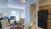 This two-story residence offers a thoughtfully designed layout that blends comfort and functionality across both levels. The main floor showcases an open-concept living and dining area bathed in natural light from large windows dressed with sheer curtains, creating a bright and welcoming ambiance. Light hardwood flooring flows throughout, adding warmth and continuity to the space.

The dining area is generously sized, comfortably accommodating a full dining set, and transitions seamlessly into the living room, which features ample space for relaxed seating arrangements. A framed piece of wall art adds a touch of character to the shared space. The kitchen is positioned adjacent to the dining area and is equipped with a full-size refrigerator, offering convenient access for everyday living.

Crisp white interior doors with classic paneling add a clean, timeless aesthetic throughout the main floor, while a staircase leads to the upper level, providing additional living space. The neutral wall tones and consistent trim work create a cohesive and polished interior that feels both spacious and inviting. The overall design strikes a balance between practical living and an appealing, well-maintained aesthetic suited for everyday comfort.