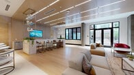 This single-floor amenity space is designed with a contemporary aesthetic, blending warm and neutral tones throughout to create an inviting, polished atmosphere. The open layout is anchored by light hardwood flooring that transitions seamlessly between distinct functional zones, while linear recessed lighting in the paneled ceiling provides consistent, even illumination across the space.

The lounge area centers around a large modular sectional sofa and accent seating, including a bold red chair that adds a pop of color against the otherwise soft palette. A striking large-format video wall, mounted against light wood paneling, serves as the focal point of this zone. Floor-to-ceiling black-framed windows and glass doors flood the space with natural light and offer views of the surrounding outdoor area.

Adjacent to the lounge, a bar-height seating section features counter stools arranged along a sleek counter, complemented by pendant lighting and integrated greenery. A built-in media credenza with a wood-tone and stone-look surface anchors a secondary display area nearby.

A more intimate nook provides a semi-private seating arrangement with a round dark table, curved boucle chairs with brass-finished legs, and textured wall paneling in layered gray and gold tones, adding warmth and visual depth to the overall design.