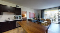 This single-floor residence offers a thoughtfully designed open-concept layout that seamlessly connects the kitchen, dining, and living areas. The kitchen is outfitted with dark espresso cabinetry, stainless steel appliances including a full-size refrigerator and electric range, light countertops, and a dishwasher. A warm wood-toned dining table anchors the dining space, which flows naturally into the living room. The living area features light hardwood flooring, a wall-mounted flat-screen TV, and access to a private balcony through sliding glass doors framed by striped curtains, offering views of surrounding greenery. The space is accented with colorful artwork and tall indoor plants throughout. The primary bedroom provides a serene retreat with plush carpeting, ample dresser storage, natural light from a window, and a wall-mounted TV. The secondary bedroom is cozy and well-lit, featuring patterned drapes and open display shelving. Two bathrooms serve the residence; one includes a tub-shower combination with tiled surround, dark vanity cabinetry, and a large mirror, while the second features a single vanity with clean white countertops and hardwood-style flooring. Overall, the space presents a modern, cohesive aesthetic with functional design throughout.