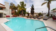 Located in Carlsbad, CA, this community outdoor amenity space at 2937–2949 Unicornio St offers a well-appointed pool area designed for relaxation and leisure. The centerpiece is a generously sized swimming pool with clear, blue-tinted water and smooth concrete decking that surrounds the entire area. Adjacent to the pool is a dedicated spa, tucked beneath a covered archway supported by stucco columns with terracotta-accented trim — an architectural detail consistent with the Spanish-influenced style of the surrounding residential buildings. The pool deck is furnished with cushioned chaise lounges for sunbathing, while a separate patio area provides multiple dining sets with tables, red chairs, and large market umbrellas for shaded outdoor seating. Lush mature trees, including tall palms and evergreens, frame the perimeter, providing privacy and a tropical ambiance. The area is enclosed by wrought iron fencing with a gated entry. A built-in barbecue grill and additional seating add to the functionality of the space. The surrounding buildings feature white stucco exteriors and red tile rooflines, complementing the overall Southern California aesthetic of this inviting outdoor community retreat.