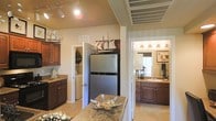 This single-floor residence is thoughtfully designed and styled with a cohesive nautical theme, reflected in decorative ship models and sailing artwork displayed throughout the space. The kitchen serves as a central hub, featuring rich dark wood cabinetry, granite countertops, and stainless steel appliances, including a full-size refrigerator. A kitchen island with matching granite provides additional workspace and a natural gathering point. Track lighting illuminates the space from above, complementing the warm, neutral tones of the walls and tile flooring. Adjacent to the kitchen, a dedicated laundry area with washer and dryer units offers convenient in-unit functionality, tucked neatly behind a doorway. The bathroom is well-appointed with dark wood vanity cabinetry, a granite-topped sink, and warm vanity lighting above a large mirror that enhances the sense of brightness. Sailing-themed artwork continues the nautical motif into this space. The overall ambiance of the home is warm and inviting, with a consistent design palette of warm beiges, dark wood tones, and stone surfaces that tie each area together seamlessly.