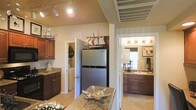 This single-floor residence is thoughtfully designed and styled with a cohesive nautical theme, reflected in decorative ship models and sailing artwork displayed throughout the space. The kitchen serves as a central hub, featuring rich dark wood cabinetry, granite countertops, and stainless steel appliances, including a full-size refrigerator. A kitchen island with matching granite provides additional workspace and a natural gathering point. Track lighting illuminates the space from above, complementing the warm, neutral tones of the walls and tile flooring. Adjacent to the kitchen, a dedicated laundry area with washer and dryer units offers convenient in-unit functionality, tucked neatly behind a doorway. The bathroom is well-appointed with dark wood vanity cabinetry, a granite-topped sink, and warm vanity lighting above a large mirror that enhances the sense of brightness. Sailing-themed artwork continues the nautical motif into this space. The overall ambiance of the home is warm and inviting, with a consistent design palette of warm beiges, dark wood tones, and stone surfaces that tie each area together seamlessly.