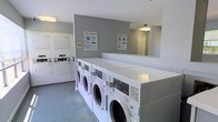 This shared laundry facility is designed with functionality and convenience in mind. The space features a clean, no-frills layout with light grey walls and a durable speckled floor surface well-suited to high-traffic use. A row of front-loading washing machines lines one side of the room, mounted on a raised white platform that provides an ergonomic working height and offers a clean, uniform appearance. Complementing these are stacked washer and dryer units positioned along the adjacent wall, maximizing the available floor space and increasing the overall laundry capacity of the facility. Instructional posters titled "Easy Washing" and "Simple Drying" are prominently displayed on the walls, offering clear guidance for users. Overhead flush-mount lighting ensures the room is well-lit throughout. A large mirror on one wall adds a sense of openness to the space. The room also includes a coin or card-operated payment station for machine activation. The overall ambiance is tidy, functional, and organized, making it a practical and accessible laundry solution for its users.