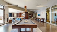 Station 40 is a thoughtfully designed communal amenity space that blends industrial character with contemporary comfort across a single expansive floor. Polished concrete flooring and rich horizontal wood-slat wall paneling establish a warm yet modern aesthetic throughout. The game room serves as a central focal point, outfitted with a full-size billiards table, a shuffleboard table, and a dedicated card and gaming table surrounded by leather chairs — all illuminated by sleek pendant and linear lighting fixtures. Adjacent lounge areas invite relaxed gatherings, featuring plush sectional sofas, accent chairs, a fireplace, wall-mounted televisions, and curated artwork. A stylish kitchenette bar anchors the social hub of the space, complete with a light quartz-topped island, bar seating, wood cabinetry, stainless steel appliances, and a striking tiled backsplash beneath the bold "Station 40" marquee signage. A cozy banquette nook with built-in seating and a wall-mounted TV offers a more intimate setting. A large roll-up garage door opens directly onto an outdoor patio furnished with seating and shade umbrellas, seamlessly connecting interior and exterior entertaining areas and allowing abundant natural light to pour in.