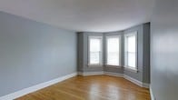 This single-floor space combines classic architectural character with a clean, understated aesthetic. The main living area immediately draws attention with its striking bay window configuration, where three white-trimmed double-hung windows curve outward to invite generous amounts of natural light throughout the room. The warm hardwood flooring runs throughout the space, adding richness and a timeless quality to the interior. Walls are finished in a soft, cool gray tone that complements both the bright white trim and the natural wood underfoot, creating a cohesive and calming palette. Crisp white baseboards provide a clean contrast along the perimeter of the room, reinforcing the traditional detailing found throughout. The ceiling features a subtle textured finish and is painted white, contributing to the overall sense of openness and airiness. The layout is straightforward and versatile, offering an open canvas well-suited to a variety of furnishing arrangements. The combination of natural light from the bay window, warm flooring, and neutral walls gives the space a welcoming ambiance that balances classic charm with modern simplicity.