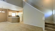 This two-story space showcases an open-concept design that seamlessly connects the main living area to the kitchen. The living space is anchored by striking vaulted ceilings that rise dramatically overhead, lending a sense of spaciousness and volume to the interior. Soft, neutral-toned carpet covers the living area floor, contributing to a warm and comfortable atmosphere throughout.

The kitchen is visible from the main living area and features white cabinetry, a white refrigerator, and a distinctive curved breakfast bar with a contrasting warm brown finish. This bar area serves as a natural dividing element between the kitchen and living space while maintaining the open feel of the floor plan. A window above the kitchen sink allows natural light to filter into the space.

The walls are finished in a neutral warm beige tone, complementing the light gray upper-level wall and the clean white trim and baseboards throughout. Recessed lighting is installed in the kitchen ceiling, providing functional illumination. A visible HVAC vent near the upper level suggests efficient climate control integrated into the architecture. Overall, the space presents a clean, functional, and airy environment suited for everyday living.