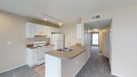 Located at 4426 Calderwood Dr in Columbus, Ohio, this single-floor unit offers a practical and well-organized layout designed for comfortable everyday living. The kitchen serves as a central hub of the space, featuring white cabinetry with gold hardware, white appliances including a range and full-size refrigerator, and a peninsula with a built-in stainless steel sink and laminate countertops. Track lighting overhead ensures the kitchen is well-lit and functional. The peninsula creates a natural transition between the kitchen and the adjacent living areas, providing both additional workspace and a casual dining or gathering spot. Tile flooring anchors the kitchen, while plush carpeting extends through the hallway and surrounding rooms, adding warmth underfoot. A hallway visible from the kitchen leads to additional rooms, with a door at the far end allowing natural light to filter into the space. Neutral wall tones throughout create a clean, versatile backdrop. Ceiling vents indicate a central HVAC system for year-round comfort. The overall layout is straightforward and efficient, making it easy to navigate and personalize.