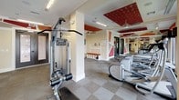 This fitness center offers a comprehensive workout environment thoughtfully designed for both cardio and strength training. The space is organized into distinct workout zones, beginning with a row of Precor cardio machines — including treadmills and ellipticals — positioned near large windows that allow ample natural light and outdoor views to energize workouts. Multiple flat-screen TVs are mounted throughout, providing entertainment during exercise sessions.

The strength training area features a variety of Precor cable machines, a functional trainer, and a dedicated free weights section with a full dumbbell rack, catering to a wide range of fitness routines. Ceiling fans are installed throughout to maintain airflow and comfort during intense training sessions.

A standout design element is the bold red patterned ceiling panels, which add a striking visual contrast to the neutral cream-toned walls and tile-patterned flooring. Geometric red and orange accent designs on the upper walls further enhance the energetic ambiance of the space. Full-length wall mirrors expand the sense of space and allow users to monitor their form. A built-in bench with storage cubbies offers a convenient spot for personal items. The overall design balances functionality with a vibrant, motivating aesthetic.