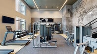 Located at 2811 S Cooper St in Arlington, Texas, this two-story fitness center is a well-appointed facility designed to support a full range of workout routines. The main floor commands attention with soaring double-height ceilings, floor-to-ceiling windows that flood the space with natural light, and views of the surrounding exterior. This level houses an extensive strength training zone, complete with cable machines, free weights, benches, and a dedicated dumbbell rack wall lined with large mirrors and mounted flat-screen TVs for an energized workout environment. A textured accent wall adds an industrial aesthetic that complements the modern equipment layout. A sculptural staircase connects the two floors and is framed by a bold, large-scale mural depicting football players in dynamic motion — a standout artistic feature that gives the facility a distinctly athletic character. The upper floor offers a dedicated cardio and group training studio with warm hardwood flooring, additional sports-themed wall murals, full-length mirrors, and a lineup of cycling bikes, rowing machines, stair climbers, and treadmills. Recessed and linear LED lighting throughout both levels ensures a bright, motivating atmosphere, making this fitness center a comprehensive and visually compelling space for residents and users alike.