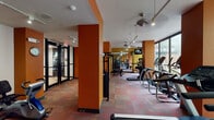 This fitness center in Houston, TX presents a spacious, single-floor layout designed to accommodate a comprehensive workout experience. The interior is defined by bold burnt-orange and mustard-yellow accent columns and walls, creating an energizing atmosphere throughout the space. Multicolored carpet tiles in warm red and earthy tones cover the floor, adding texture and visual interest underfoot.

The facility is generously outfitted with a wide range of equipment. A dedicated cardio zone features a row of treadmills along one wall, complemented by multiple elliptical trainers positioned near the large floor-to-ceiling windows. Strength training is well-represented with cable machines, weight benches, a leg press, free weights, and medicine balls distributed across the space.

Large glass windows along the perimeter flood the interior with natural light and offer views of the surrounding outdoor courtyard and neighboring architecture, enhancing the open feel of the space. Ceiling fans and track lighting provide functional illumination and airflow throughout.

A wall-mounted television is available for entertainment during workouts. The entrance features glass doors with clearly marked exit signage, and a vending machine is visible near the entry area. Overall, the fitness center is practical, well-lit, and thoughtfully organized for efficient use.