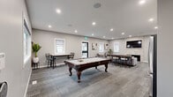 Located at 5501 Prefontaine Ave in Regina, Saskatchewan, this well-appointed clubhouse offers a stylish and versatile communal space designed for both relaxation and recreation. The open-concept layout is finished with neutral grey walls, recessed pot lighting, and patterned carpet flooring that ties the space together cohesively.

The games area anchors one side of the clubhouse, featuring a full-sized pool table as its centrepiece, complemented by bar-height tables and stools — ideal for casual socializing and friendly competition. Multiple windows throughout the space allow natural light to filter in, creating a bright and welcoming atmosphere.

The lounge area offers a refined seating arrangement with a sectional sofa and modern accent chairs gathered around a sleek linear fireplace set into a striking stacked stone feature wall. A wall-mounted flat-screen television above the fireplace adds a functional entertainment element to the space.

Toward the rear, a compact kitchenette provides added convenience, outfitted with white upper cabinetry, dark lower cabinets, a stainless steel refrigerator, and a sink with counter space. Washroom facilities are also accessible from this area. The overall design is clean and contemporary, making this clubhouse a practical and attractive amenity for the community it serves.