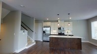 This well-appointed two-story home showcases a thoughtfully designed interior that blends modern aesthetics with functional living. The main level features an open-concept layout anchored by a stylish kitchen, where crisp white shaker cabinetry reaches toward the ceiling, complemented by a classic white subway tile backsplash. A substantial kitchen island with a rich, dark wood base and light countertop serves as both a prep space and a natural gathering point, fitted with a deep sink. Stainless steel appliances, including a French door refrigerator with ice and water dispenser, a built-in microwave, and a gas range, complete the culinary space. Pendant lighting with warm amber glass shades hang above the island, adding a refined touch to the kitchen's ambiance. Recessed lighting throughout ensures the space is well-lit. Rich, dark hardwood floors run across the main level, providing a striking contrast to the soft, neutral gray walls. A staircase with a dark wood handrail and white balusters leads to the second floor, hinting at the additional living space above. The overall atmosphere is clean, contemporary, and move-in ready.