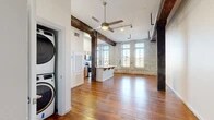 Located at 301 Crockett St in downtown Shreveport, Louisiana, this 2-bedroom, 1-bathroom loft blends historic industrial character with modern updates. The open-concept living and kitchen area is defined by original hardwood floors, exposed heavy timber beams, and whitewashed brick walls that speak to the building's storied past. Tall arched windows line the living space, flooding the interior with natural light and offering views of the surrounding cityscape. The kitchen features stainless steel appliances, marble-style countertops, warm wood cabinetry, and a peninsula with bar seating — seamlessly connecting to the living area. A stacked washer and dryer unit is conveniently tucked near the entrance. The two bedrooms carry the same rich hardwood flooring and exposed brick accents, with the primary bedroom offering generous proportions and natural light through large windows. The bathroom is clean and functional, equipped with a full tub-shower combo and wood vanity. Track lighting and a ceiling fan complement the industrial aesthetic throughout. This loft offers a distinctive urban living experience rooted in authentic architectural detail.