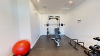 Located in San Pedro, CA, this dedicated fitness center offers a well-equipped workout space designed for effective strength and functional training. The room features crisp white walls, recessed lighting throughout, and durable rubber flooring that provides both safety and comfort underfoot.

The space is outfitted with a professional-grade Hammer Strength power rack paired with a adjustable weight bench and Olympic plates, making it ideal for a full range of strength training exercises. A Life Fitness functional trainer offers cable-based workouts and full-body conditioning options. A tiered dumbbell rack stocked with a comprehensive set of free weights lines one wall, complemented by a kettlebell and a stability ball for added versatility.

Large floor-to-ceiling windows and a glass-paneled door at one end of the room allow natural light to fill the space, while also providing direct access to an outdoor patio area, seamlessly connecting indoor and outdoor environments. The clean, minimalist aesthetic of the white walls and modern equipment creates a focused and motivating workout atmosphere. Adequate ventilation is provided via ceiling-mounted HVAC vents, ensuring comfort during intense training sessions. This fitness center delivers a comprehensive and well-appointed exercise environment suited to a variety of fitness routines.