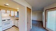 This single-floor unit offers a straightforward, functional layout spread across several rooms. The living area is generously sized, featuring plush wall-to-wall carpeting in a neutral gray tone, warm wood baseboards, and windows fitted with blinds that allow natural light to filter through. A door leads directly outside, enhancing accessibility and ventilation.

The kitchen is positioned adjacent to the living space, separated by a half-wall with a pass-through counter. It features white cabinetry with warm wood accents, light countertops, a double sink, and a white gas range with an overhead exhaust hood, providing a complete and practical cooking environment. The kitchen floor is finished with tile.

Multiple bedrooms are present throughout the unit, each carpeted and finished with neutral walls and wood baseboards. The bedrooms receive natural light through individual windows. At least one bedroom includes a walk-in closet with built-in shelving and hanging rods, offering ample storage. Additional doors throughout suggest closet or utility storage access.

Overall, the space maintains a clean, understated aesthetic with consistent neutral tones, carpet flooring in living and sleeping areas, and a practical design that prioritizes functionality and ease of use.