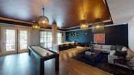 This single-floor amenity space is thoughtfully designed to accommodate both social gatherings and relaxed lounging, divided into distinct yet interconnected zones. The game room, clearly marked at its entrance, anchors one side of the space and features a full shuffleboard table as its centerpiece, complemented by rich wood-toned flooring and dramatic woven pendant light fixtures suspended from a warm, wood-paneled ceiling. Deep teal accent walls and bold artwork add a vibrant, contemporary character throughout.

Adjacent to the game room, a bar and dining area offers a striking yellow countertop island with a dark marble base, surrounded by leather-toned bar stools. Teal cabinetry with open metal shelving lines the back wall, and large floor-to-ceiling windows with sheer curtains allow generous natural light and views to the outdoors.

A separate lounge area provides a quieter retreat, anchored by a sleek black feature wall housing both a mounted television and a contemporary gas fireplace. Comfortable seating arrangements, a modern globe chandelier with brass accents, and warm hardwood flooring create an inviting and sophisticated atmosphere throughout this multifunctional communal space.