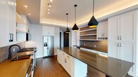 This single-floor residence showcases a thoughtfully designed open layout with a cohesive blend of contemporary finishes and warm materials. The kitchen serves as a centerpiece of the space, featuring crisp white shaker cabinetry with matte black hardware, dark stone countertops, and a generously sized center island illuminated by striking geometric black pendant lights with gold interiors. A textured tile backsplash adds visual depth, while stainless steel appliances—including a gas range, built-in microwave, and French door refrigerator—complete the culinary setup. Open shelving and a built-in bar area provide both function and style. Rich hardwood flooring flows seamlessly throughout the entire residence, grounding each area with warmth. The living area is defined by expansive floor-to-ceiling windows framed in dark metal, flooding the space with natural light and offering open urban views, with access to a private balcony. Additional rooms feature large windows with similar city outlooks, maintaining the airy, elevated feel. The bathroom is a refined retreat, boasting a dual-sink vanity with dark stone counters, framed mirrors, pendant lighting, large-format tile flooring, and a glass-enclosed shower with warm wood-toned tile walls.