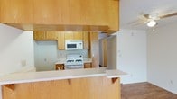 This single-floor space is designed with practicality and an open feel in mind. The kitchen serves as a central feature, outfitted with warm honey-toned wood cabinetry that extends from floor to ceiling, providing generous storage throughout. A gas range and built-in microwave are positioned along the back wall, complemented by light-colored countertops that create a clean, bright workspace. A peninsula with a breakfast bar extends outward, offering additional prep space and casual seating while visually connecting the kitchen to the adjoining living area. The pass-through opening above the peninsula enhances the open-concept feel, allowing natural sightlines between the kitchen and the rest of the space. The living area features carpeted flooring, offering a softer contrast to the kitchen's hard surfaces. A hallway visible from the kitchen leads further into the unit, suggesting additional rooms beyond. The textured ceiling is consistent throughout, adding a classic residential character to the space. Overall, the layout emphasizes connectivity and efficient use of space, making it well-suited for everyday living and easy entertaining.