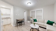 This single-floor unit offers a practical and cohesive layout designed for comfortable everyday living. The interior is unified by light hardwood-style flooring and neutral white and grey tones, creating a clean, bright atmosphere throughout. Textured ceilings add a subtle architectural detail consistent across all rooms.

The living area accommodates both lounging and dining, with a window dressed in horizontal blinds allowing natural light to filter in. A radiator beneath the window provides heating, and there is adequate space for a sofa, coffee table, and a small dining set. A doorway leads further into the unit from this space.

A hallway connects the main living area to the remaining rooms, with the kitchen visible at the far end. The kitchen features white cabinetry, a sink, and a full-size refrigerator, offering essential functionality in a compact footprint.

Two bedrooms are accessible from the hallway. Both rooms share a similar aesthetic, with light walls, textured finishes, and windows fitted with blinds that admit natural light. Each room comfortably fits a bed and nightstand. Radiator units are present in the bedrooms, providing consistent heating.

The bathroom is straightforward and clean, featuring a soaking tub with a shower rod, a toilet, and light flooring that matches the rest of the unit.