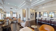 This elegantly designed single-floor amenity space makes a strong first impression with its grand entry, framed by dark-framed glass double doors, coffered ceilings, and a striking pendant chandelier above a marble-topped entry table adorned with fresh orchids. Dark hardwood floors flow seamlessly throughout, anchoring the warm, neutral color palette of taupe, cream, and amber tones.

The open-plan layout encompasses multiple distinct seating areas furnished with plush sofas, textured lounge chairs, and accent seating in rich cognac leather, all arranged around polished stone side tables and soft area rugs. Tray ceilings with recessed lighting add architectural depth and a refined sense of volume to each zone.

A long, sleek bar counter with white upholstered bar stools and pendant lighting serves as a central social hub, complemented by a dark wood accent wall that adds warmth and contrast. Framed artwork and tall white decorative vessels with organic branch arrangements punctuate the space with curated style.

Floor-to-ceiling windows line the perimeter, flooding the interior with natural light and offering expansive views of the surrounding skyline and landscape. Sheer layered drapery softens the light while maintaining an airy, sophisticated ambiance throughout this impressive communal space.