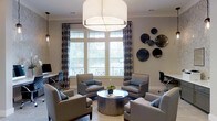 This well-appointed single-floor space exudes a refined, contemporary ambiance designed for comfort and social interaction. The focal point of the lounge area is a large, cylindrical drum pendant light that anchors the seating arrangement below, casting a warm and inviting glow throughout the room. Natural light pours in through generously sized windows dressed with stylish patterned curtains, offering views to the outdoors and creating a bright, open atmosphere. The seating area is thoughtfully arranged with a set of plush upholstered armchairs in a neutral gray tone, centered around a sleek, dark circular coffee table — an ideal configuration for conversation and gathering. The walls feature a tasteful display of decorative dark metallic disc accents, adding an artistic and modern touch to the overall aesthetic. Crown molding along the ceiling contributes a classic architectural detail, while the light-toned walls and flooring provide a clean, versatile backdrop. A workspace with a desk and chair is also visible, suggesting the space serves a dual purpose for both relaxation and productivity. Overall, the space strikes a balance between functionality and sophisticated design.