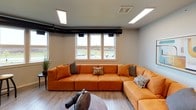 This single-floor amenity space is thoughtfully designed to offer a welcoming and versatile environment for socializing and recreation. The layout is divided into distinct functional zones, each with its own character while maintaining a cohesive contemporary aesthetic throughout.

The lounge area serves as a central gathering point, anchored by a generous L-shaped sectional sofa in a bold burnt orange upholstery that adds a vibrant pop of color to the neutral palette. Large windows line the walls, flooding the space with natural light and offering views of the surrounding outdoor greenery. Light wood-look flooring runs throughout, complementing the clean, modern feel.

Adjacent to the lounge is a dedicated entertainment and game room, featuring a wall-mounted flat-screen television, a media console, and a Golden Tee arcade game — ideal for casual recreation. The gray accent walls and recessed LED panel lighting give this zone an energetic yet polished ambiance.

The kitchenette is well-appointed with rich dark wood cabinetry, granite-style countertops, a double sink, and a full-size stainless steel refrigerator. A classic white subway tile backsplash adds a clean contrast, while a curated arrangement of woven decorative plates on the wall introduces a warm, artistic touch. Together, these spaces create a dynamic and inviting community environment.