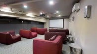 This purpose-built screening room delivers a true cinematic experience within a single-floor layout. At its focal point is a large wall-mounted projection screen paired with a ceiling-mounted projector, creating a dedicated viewing setup designed for immersive entertainment. The seating arrangement features multiple deep red modular sofas and lounge chairs positioned in a tiered-style layout, ensuring clear sightlines to the screen from every seat. The recessed ceiling lighting provides ambient illumination without interfering with the viewing experience, while exposed utility elements along the ceiling add an industrial character to the space. A wall-mounted air conditioning unit ensures comfort during extended viewing sessions. The light-toned hexagonal tile flooring contrasts attractively with the bold red upholstery, contributing to the room's distinct aesthetic. Side tables are thoughtfully placed near the seating for added convenience. The overall ambiance is intimate and focused, combining functional design with a stylish, lounge-like atmosphere that makes this space well-suited for movie screenings, sports viewing, or group entertainment gatherings.