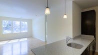 This single-floor space showcases a thoughtfully designed open-concept layout that seamlessly connects the kitchen and living areas. The kitchen features a speckled granite countertop that provides generous prep space, complemented by a stainless steel sink with a modern arc-neck faucet. Two pendant lights with warm-toned glass shades hang overhead, providing both functional task lighting and a stylish focal point above the kitchen counter.

The living area extends naturally from the kitchen, unified by light-toned hardwood flooring that runs throughout the space, contributing to a clean and cohesive aesthetic. Neutral cream-colored walls enhance the sense of openness and allow natural light to reflect freely throughout the interior. A window positioned along the far wall invites natural light into the living area, brightening the space and creating a welcoming atmosphere.

The open floor plan promotes a fluid, connected feel between functional zones, making the space well-suited for both everyday living and entertaining. The overall ambiance is light, airy, and contemporary, with a minimalist design sensibility that emphasizes clean lines and practical finishes.