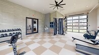 Located at 1801 Walt Whitman Dr, this fitness center offers a thoughtfully designed exercise space spread across a single level. The room is defined by its striking checkerboard-patterned flooring in neutral tan and white tones, which lends a clean, polished aesthetic throughout. Large windows line multiple walls, flooding the space with natural light and providing views of the surrounding community. A glass garage-style door on one side further enhances the connection to the outdoors, allowing for additional ventilation and an open atmosphere. Oversized ceiling fans overhead contribute to comfort during workouts. The cardio zone is well-stocked with Matrix treadmills, elliptical machines, stationary bikes, and a rowing machine. The strength training area features a Matrix functional trainer with cable attachments and resistance bands, a rack of kettlebells and medicine balls, a weight bench, and a dumbbell rack. Textured stone-look wall cladding adds an elevated design element, while a built-in shelving niche provides storage. An accessible restroom is conveniently accessible from within the space. Double glass doors connect to an adjacent amenity area, and direct exterior access is available via a keyed exit door, making this fitness center a comprehensive and inviting facility.