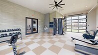 Located at 1801 Walt Whitman Dr, this fitness center offers a thoughtfully designed exercise space spread across a single level. The room is defined by its striking checkerboard-patterned flooring in neutral tan and white tones, which lends a clean, polished aesthetic throughout. Large windows line multiple walls, flooding the space with natural light and providing views of the surrounding community. A glass garage-style door on one side further enhances the connection to the outdoors, allowing for additional ventilation and an open atmosphere. Oversized ceiling fans overhead contribute to comfort during workouts. The cardio zone is well-stocked with Matrix treadmills, elliptical machines, stationary bikes, and a rowing machine. The strength training area features a Matrix functional trainer with cable attachments and resistance bands, a rack of kettlebells and medicine balls, a weight bench, and a dumbbell rack. Textured stone-look wall cladding adds an elevated design element, while a built-in shelving niche provides storage. An accessible restroom is conveniently accessible from within the space. Double glass doors connect to an adjacent amenity area, and direct exterior access is available via a keyed exit door, making this fitness center a comprehensive and inviting facility.